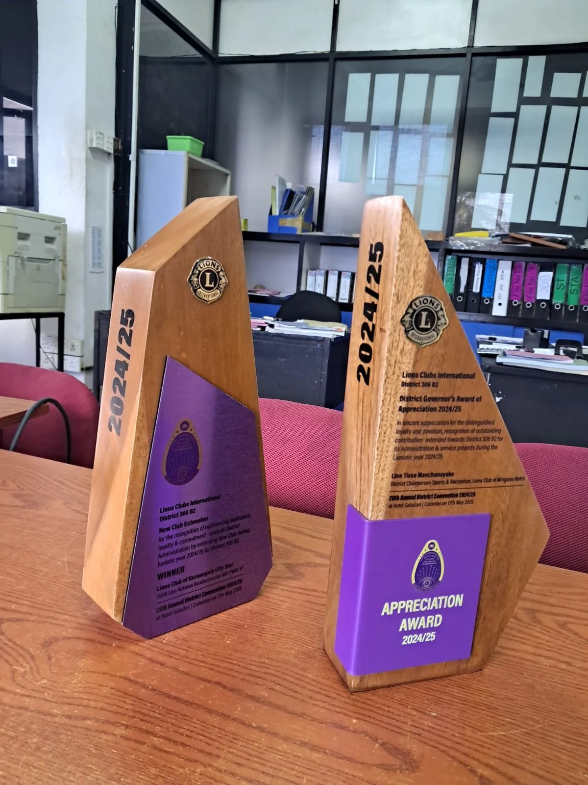 Two wooden trophies with Lions Clubs International logos and purple plaques, placed on a wooden table in an office setting.