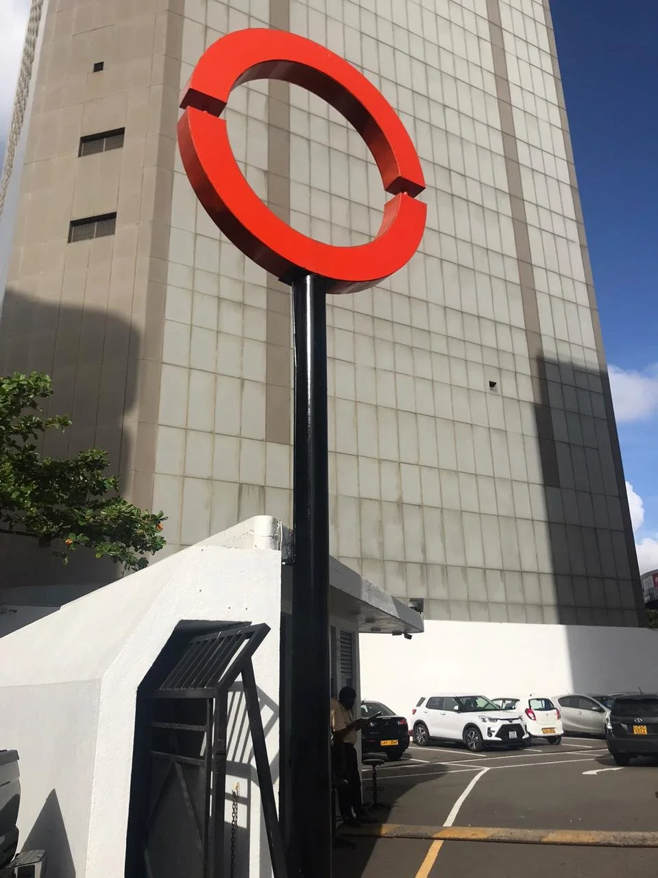 A large red circular sign on a black pole in front of a tall beige building with small windows and shadowed areas, with parked cars and a person standing near a bench in the background.