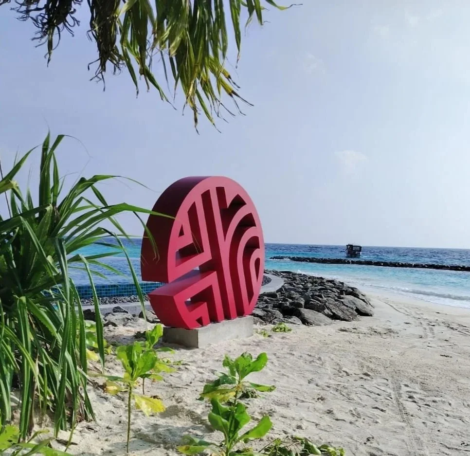 A pink, round sculpture with intricate cut-out patterns is on a sandy beach near the ocean, with green plants in the foreground and a clear sky overhead.