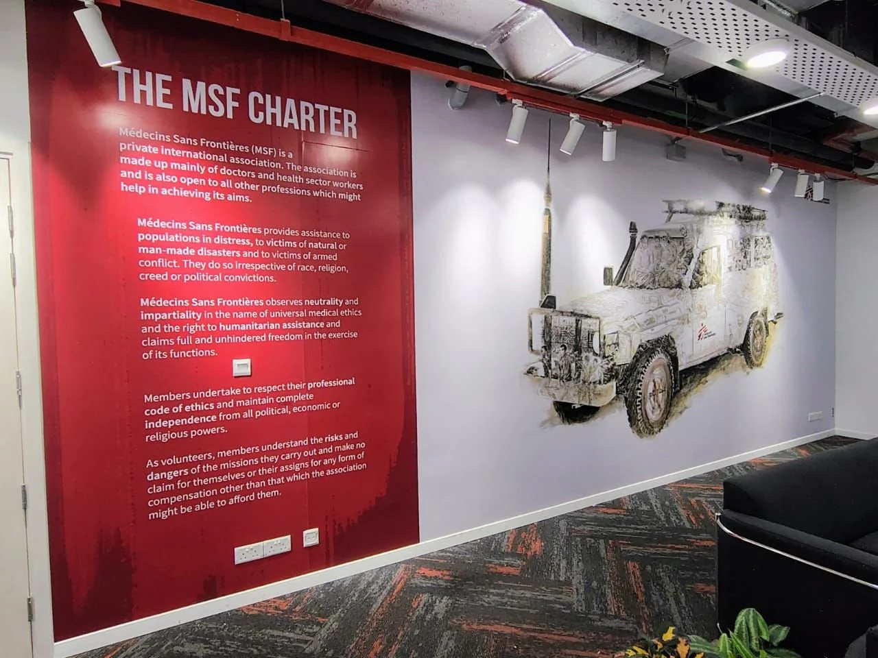 A wall display with red background text explaining the MSF charter on the left and an illustration of a white, rugged vehicle on the right, with a mural of the Eiffel Tower in the background.
