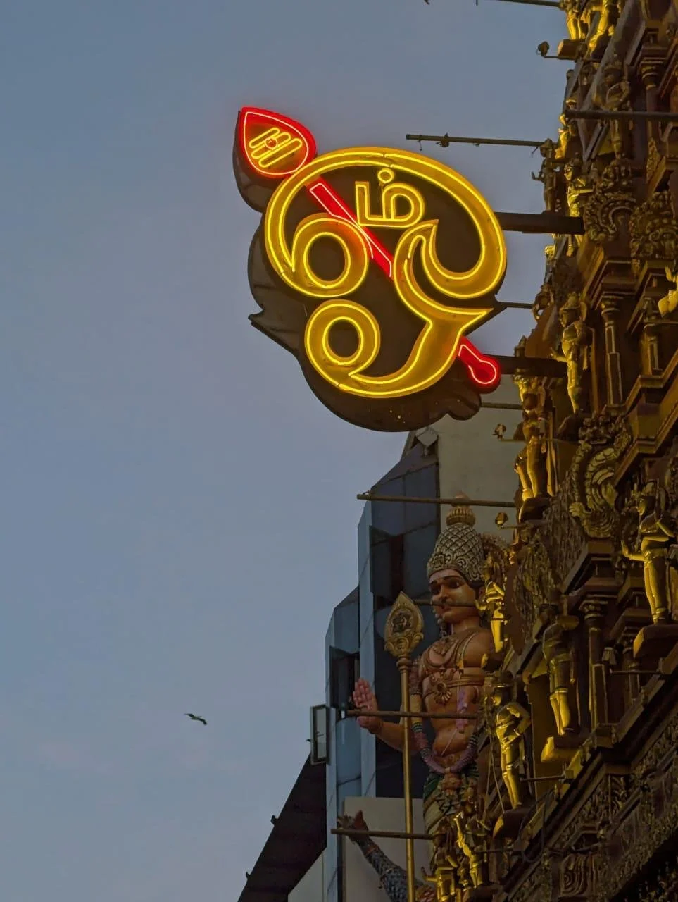 Neon sign with Hindi text, mounted on an ornately decorated building with statues, during twilight.