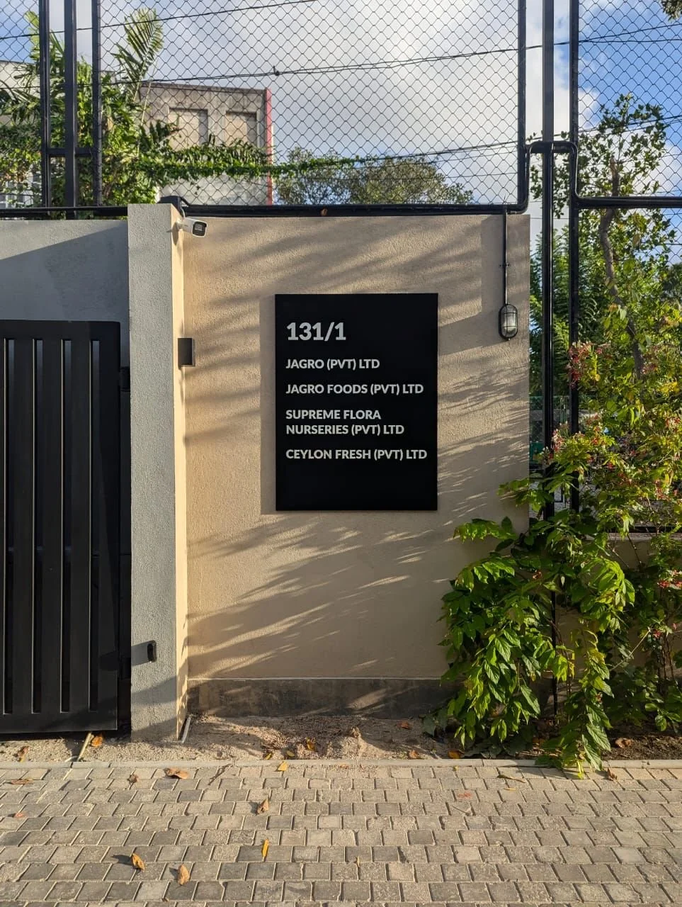 Building entrance sign displaying company names at 131/1, including Jagro (Pvt) Ltd, Jagro Foods (Pvt) Ltd, Supreme Flora Nurseries (Pvt) Ltd, and Ceylon Fresh (Pvt) Ltd, with a grey wall and plants nearby.