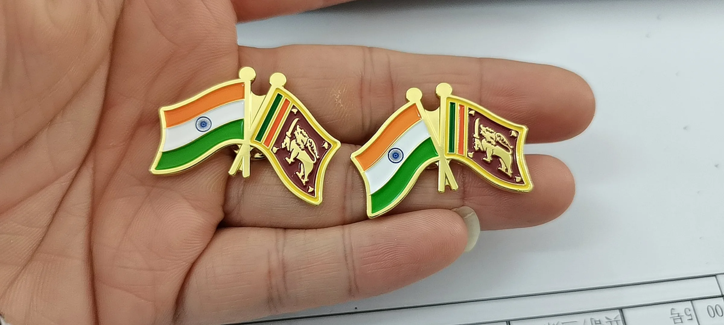 Two small lapel pins featuring the flags of India and Sri Lanka, with the Indian flag on the left and the Sri Lankan flag on the right, held on a person's fingers.