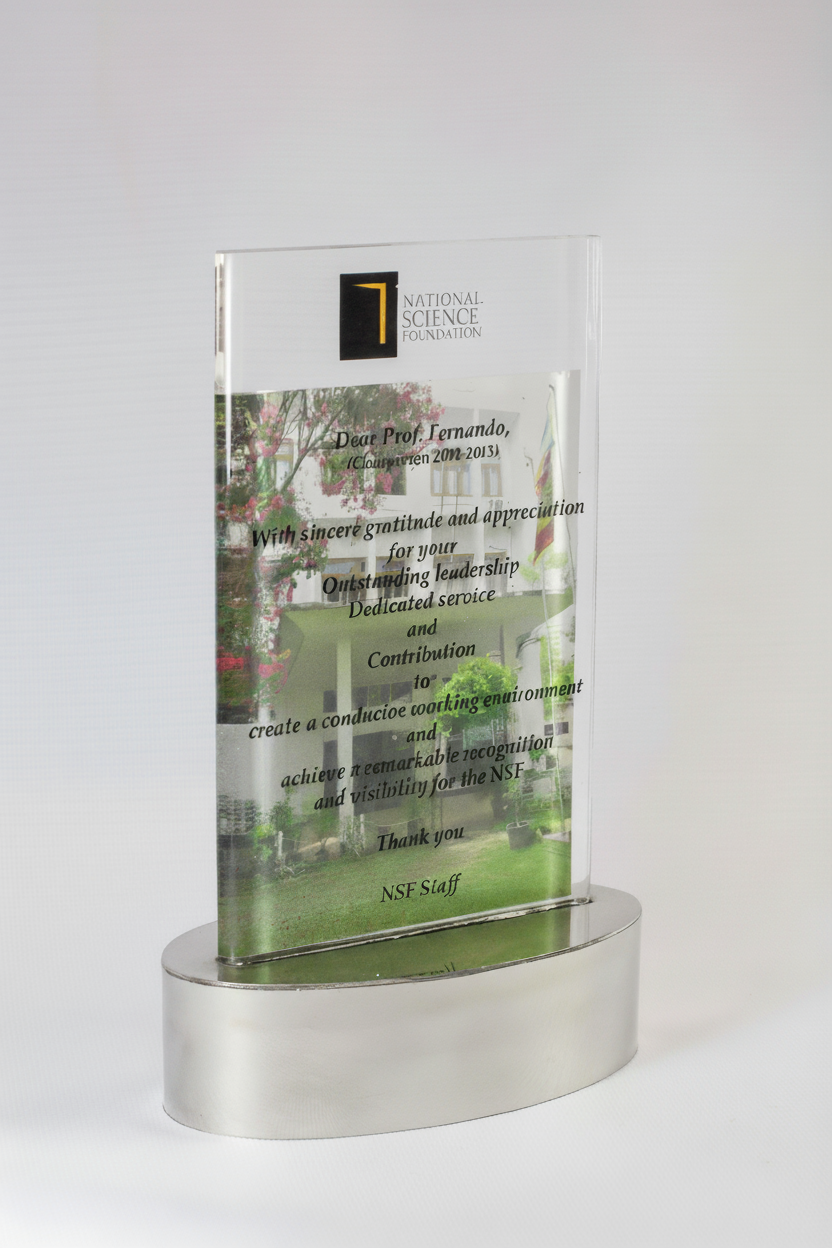 A clear glass award with a metal base, engraved with a message of appreciation from the National Science Foundation for Prof. Fernando's outstanding leadership and contribution, featuring a background image of a house and trees.