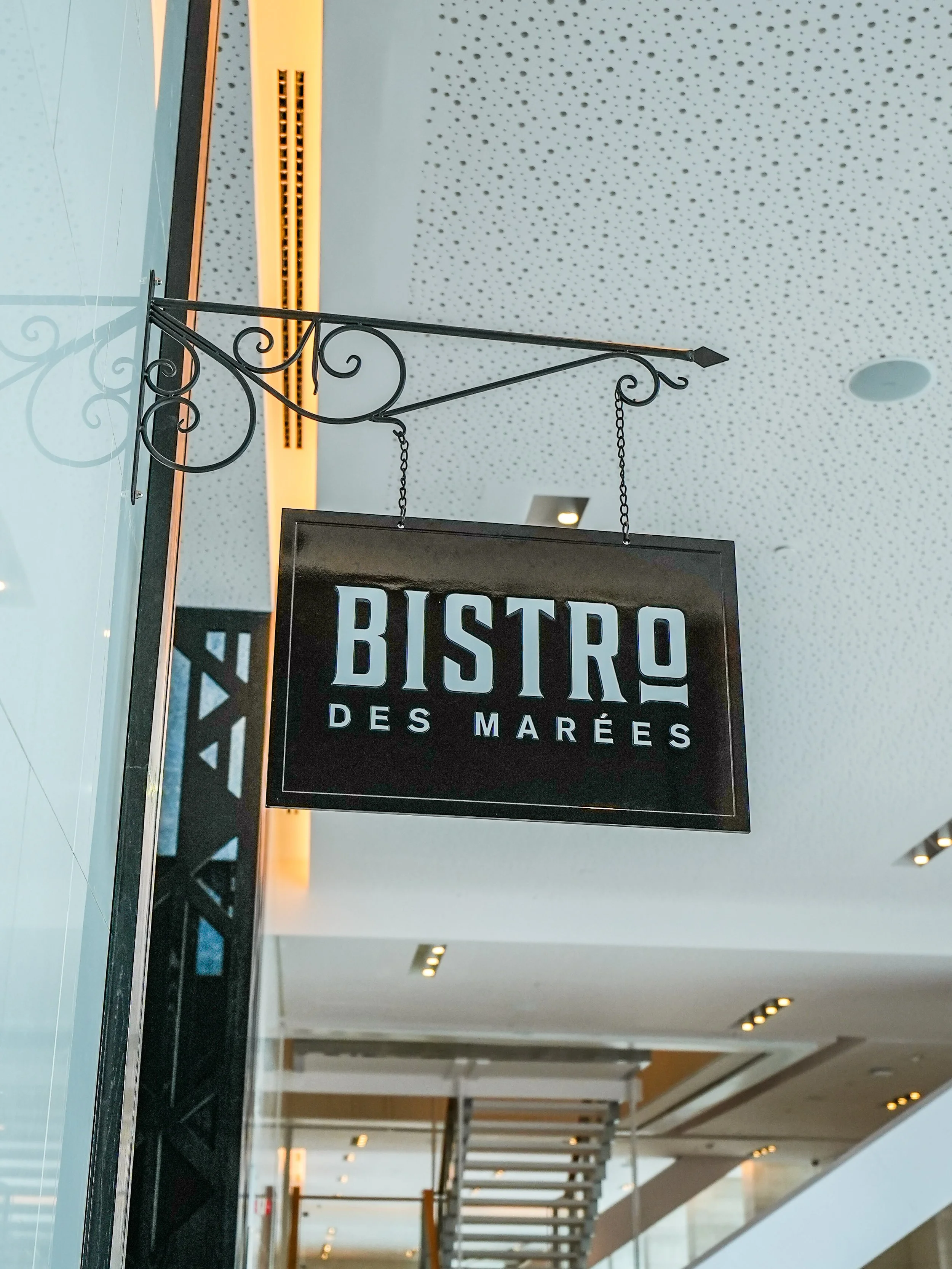 Signboard reading 'BISTRO DES MARÉES' hanging from a decorative black wrought iron bracket in a modern interior with ceiling lights and staircase.