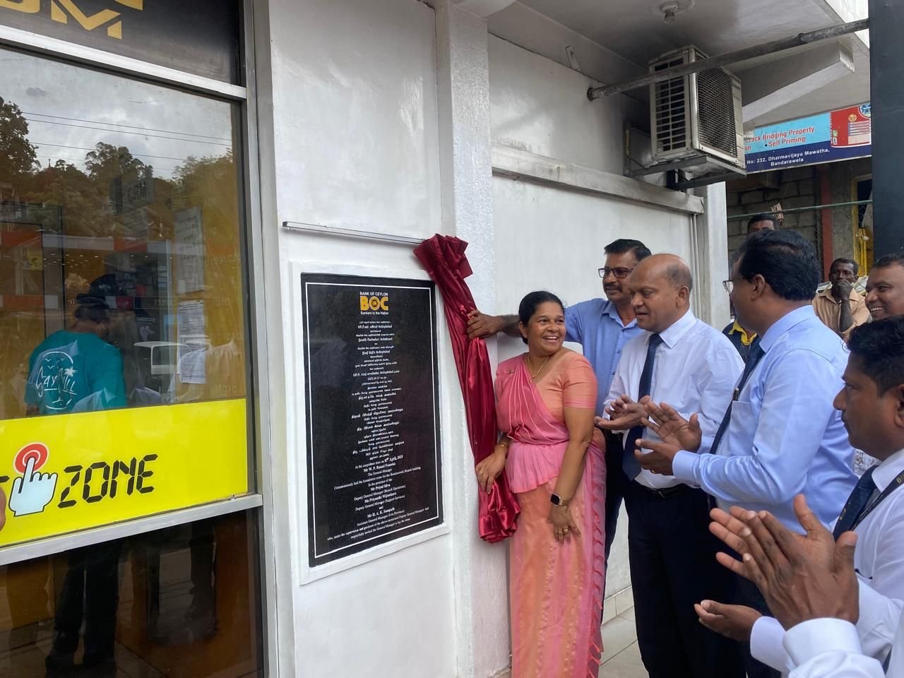 A group of officials and a woman in a pink saree are unveiling a plaque outside a building during a ceremony. The woman in the pink saree is smiling, and others are clapping and smiling.