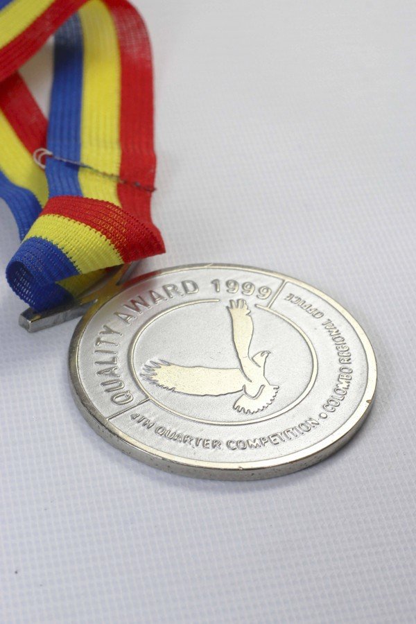 A silver medal with a colorful ribbon in red, yellow, and blue, featuring an eagle and the words "Quality Award 1999" and "Quarter Quarter Collection"
