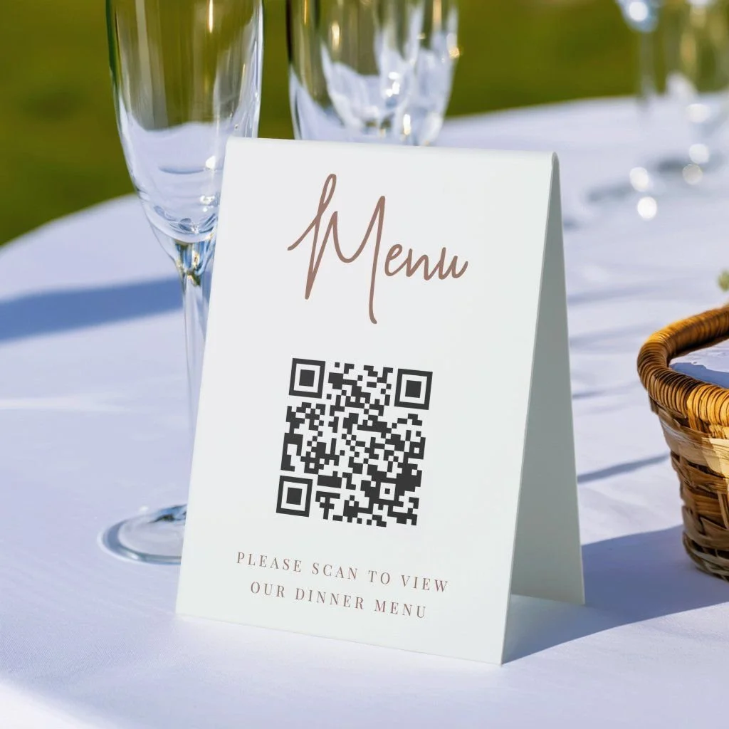 A tabletop with a white tablecloth holding a menu sign with a QR code, wine glasses, and a wicker basket, outdoors with a blurred green background.