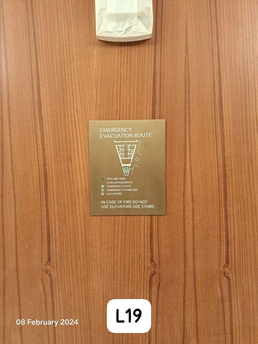 A wooden wall with an emergency evacuation route sign and a white paper bag above it. The sign shows the evacuation route map, nearby exits, staircases, and elevators, and advises not to use elevators in case of fire. The bottom of the wall has a lab