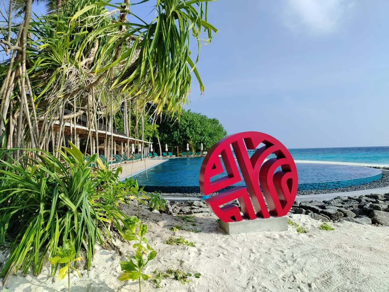 A coastal scene with a sandy beach, lush green tropical plants, a blue swimming pool, and the ocean beyond. There is a pink decorative sculpture near the pool and sun loungers with umbrellas in the background under a partly cloudy sky.