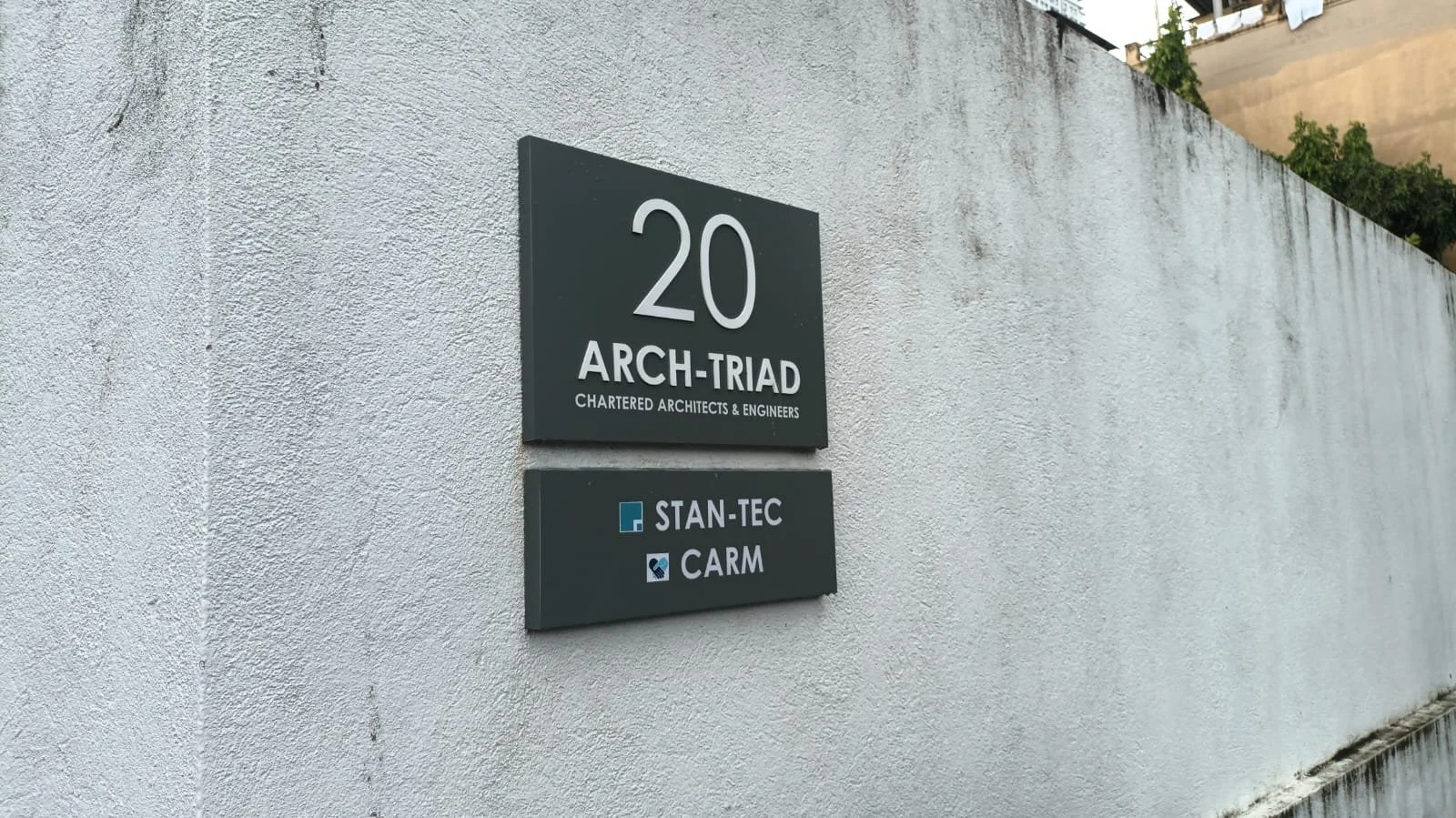 Exterior wall with two business signs above a white textured wall, with some greenery visible at the top.