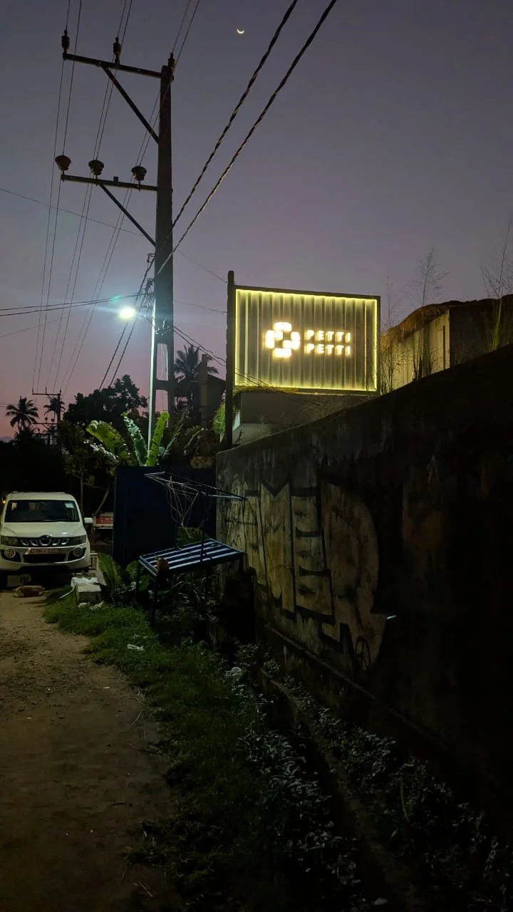 Nighttime scene with a power pole, streetlight, a car parked on a dirt road, a neon sign reading 'Petti Petti', and a crescent moon in the sky.