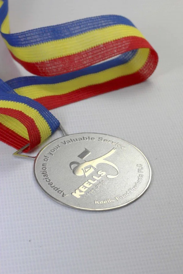 A silver medal with the inscription "Recognition of your Valuable Service" and the logo of Keells Food Primitives PLC. It is attached to a ribbon with blue, yellow, and red stripes.