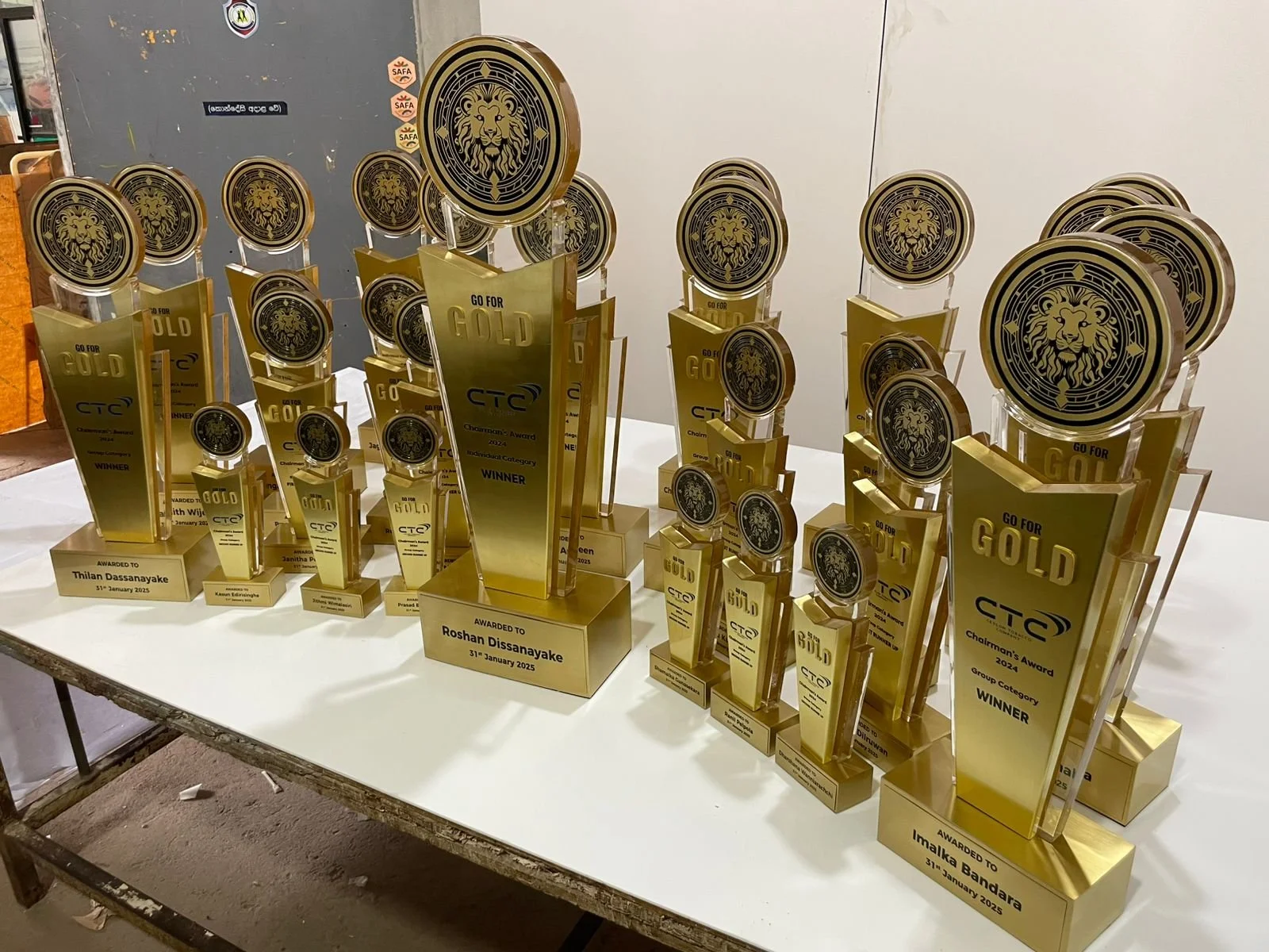Multiple gold and black trophies displayed on a white table, each labeled with the recipient's name and award details, including categories like 'Winner' and 'Chairman's Award' from the CTTC awards ceremony held on January 31, 2025.