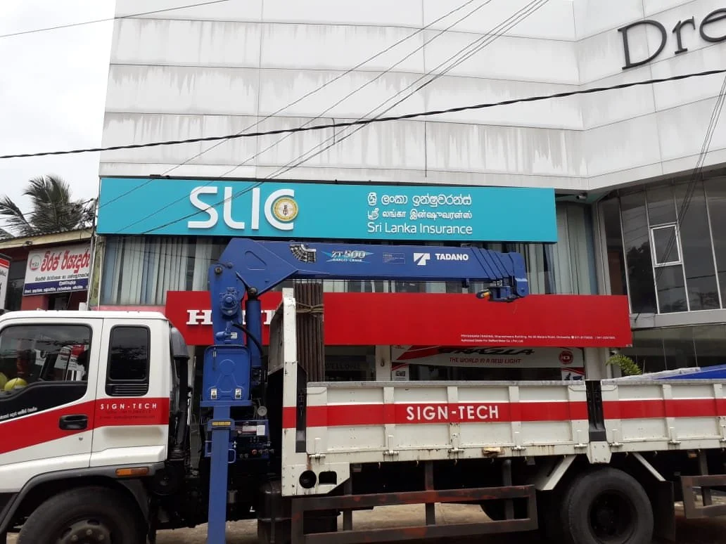 A street scene with a truck and a building in the background. The truck has equipment for installing signboards, and there are various signs, including one with the Sri Lanka Insurance logo and another for Sign-Tech.