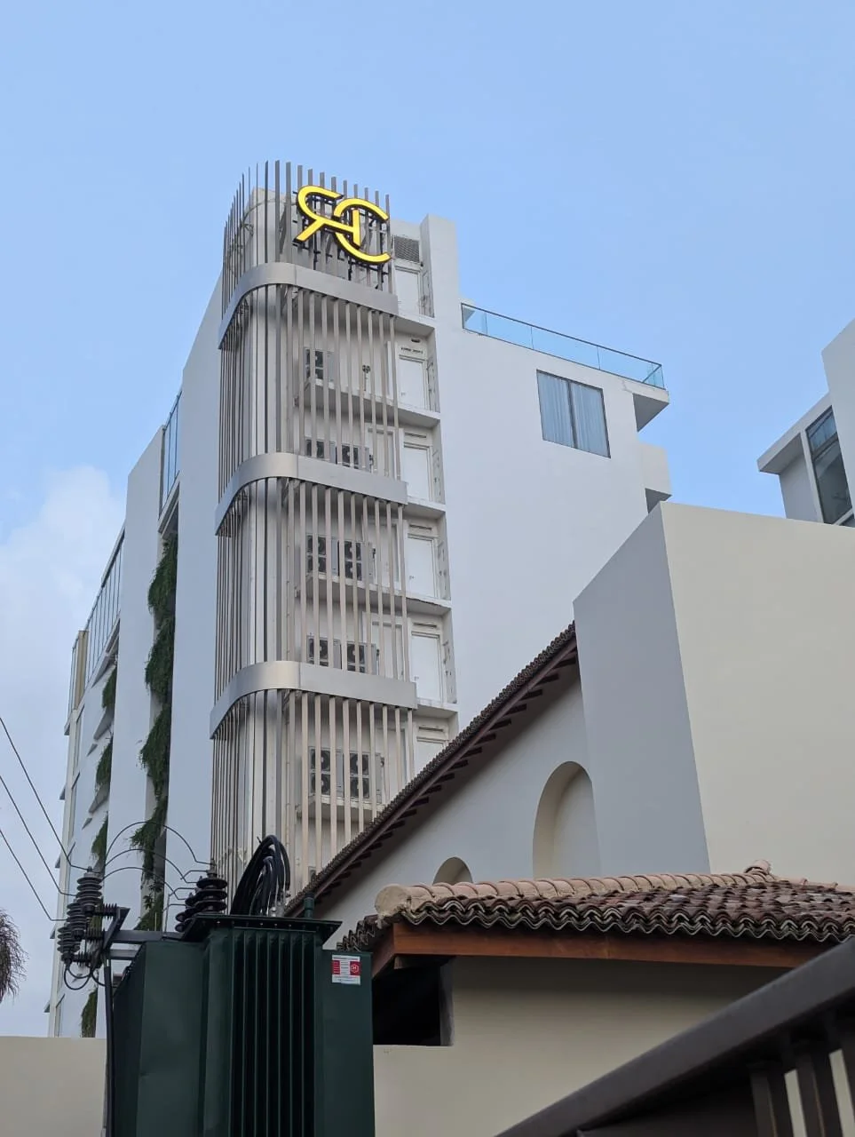 Modern white multi-story building with vertical metal slats on side, logo at top, and a tiled roof building in foreground.
