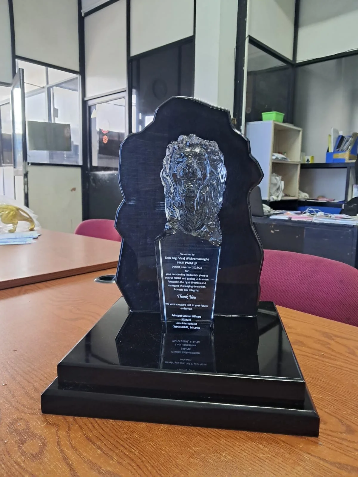 A black and clear glass lion head trophy on a black base, placed on a wooden table in an office setting.