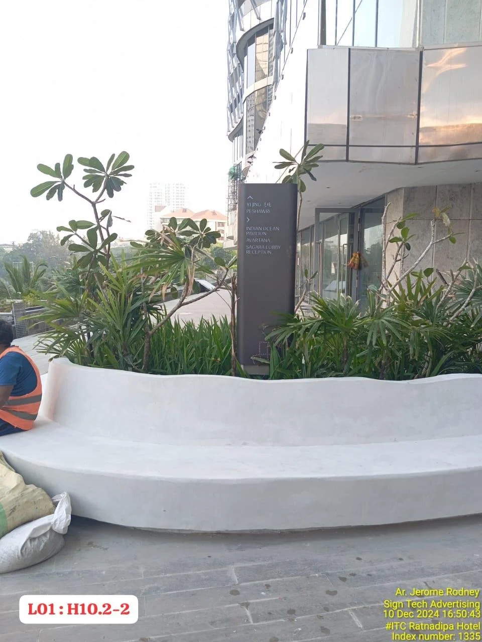 A modern building with glass windows and a concrete exterior, surrounded by green plants. There is a black sign on the building with white text. A white curved bench is in the foreground, with a person wearing a safety vest sitting beside it. There a