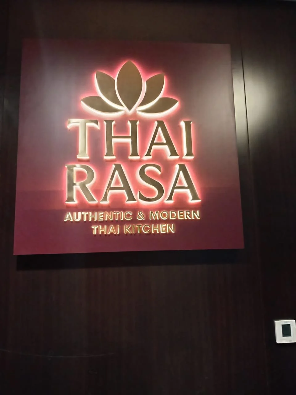 A illuminated sign featuring a lotus flower graphic with the text "THAI RASA" and the tagline "Authentic & Modern Thai Kitchen" on a dark wall.