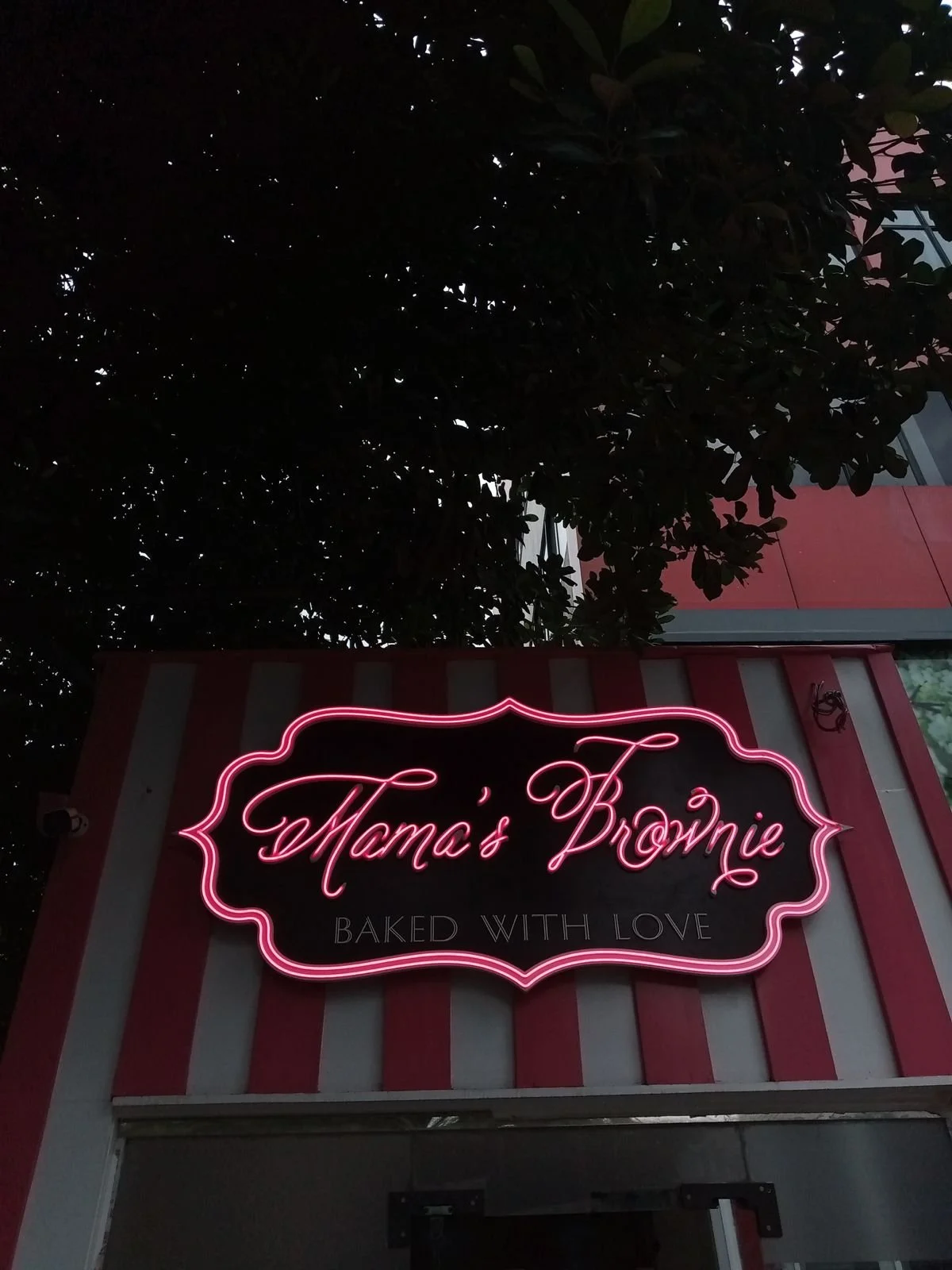 Neon sign with pink light on a bakery storefront reading: 'Mama's Brownie, Baked with Love' with red and white striped background and a tree overhead.