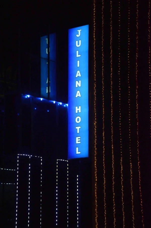 Blue vertical sign reading "JULIANA HOTEL" illuminated at night, with decorative string lights on the building.