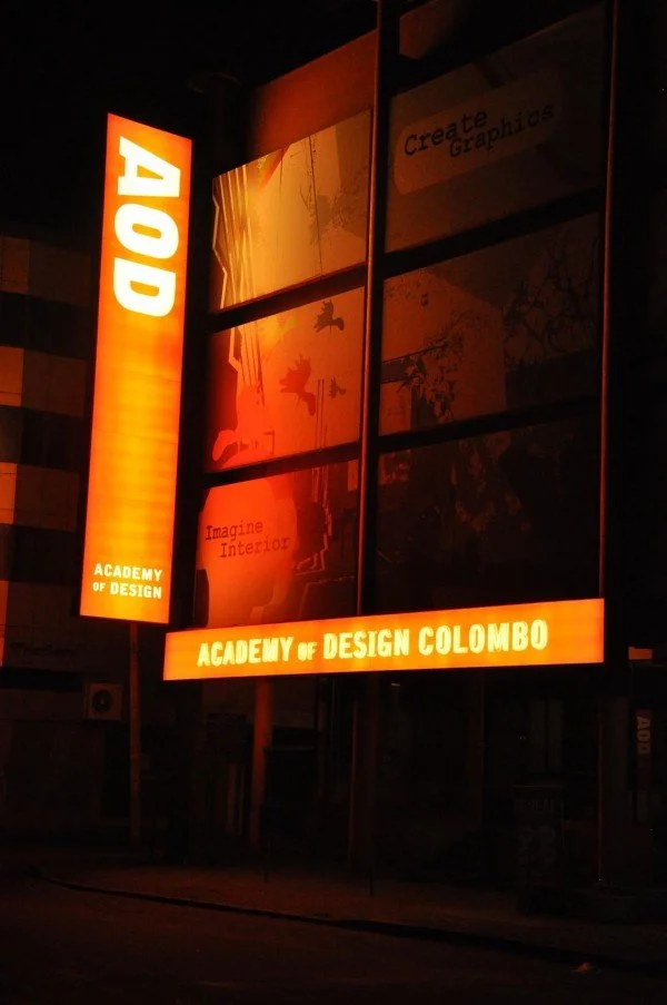Illuminated red and orange vertical signs for the Academy of Design Colombo, with graphics and text promoting design courses.