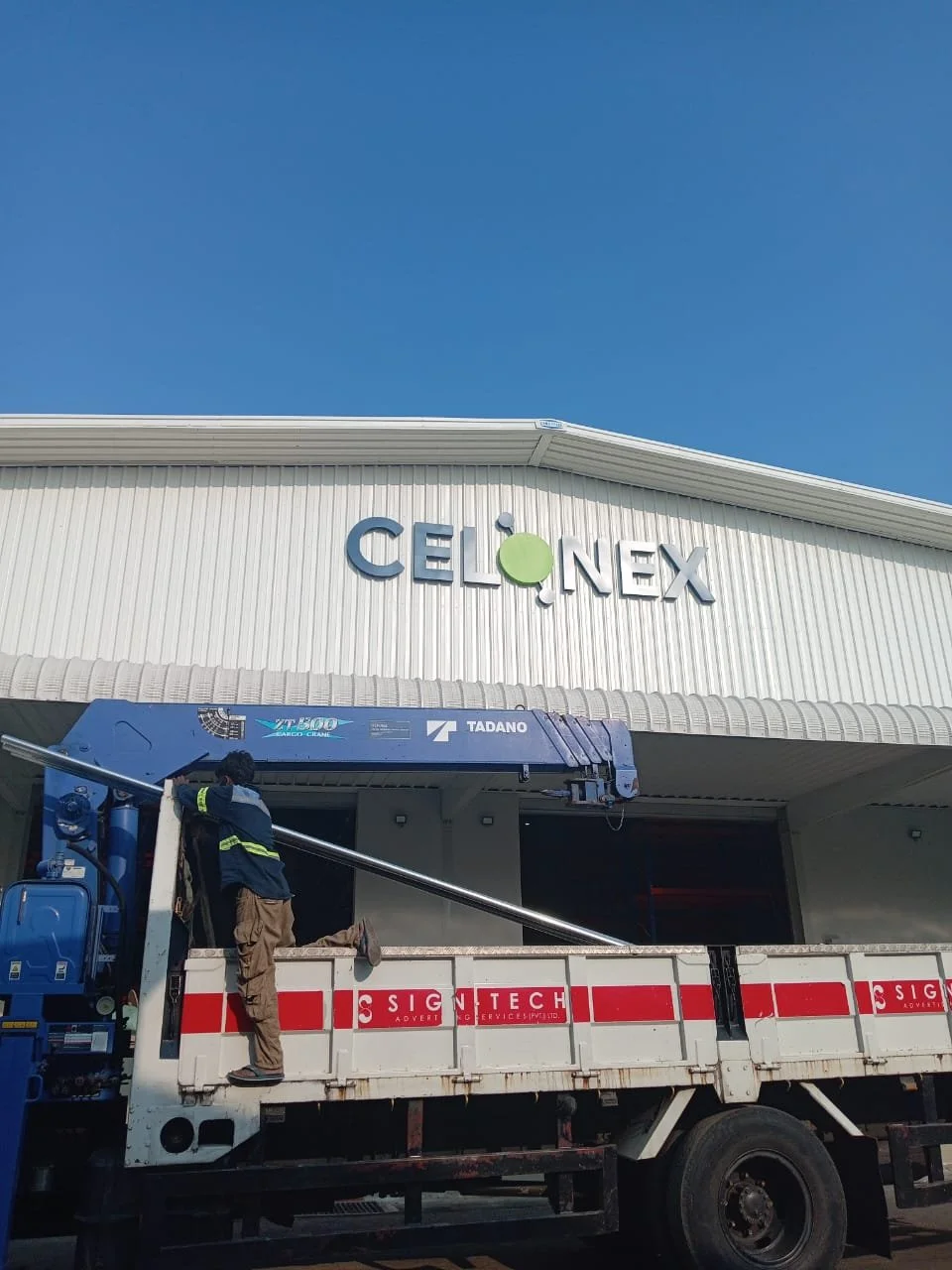 Worker on a truck installing or repairing a sign for Celonex at a building with a white exterior and a blue sky background.