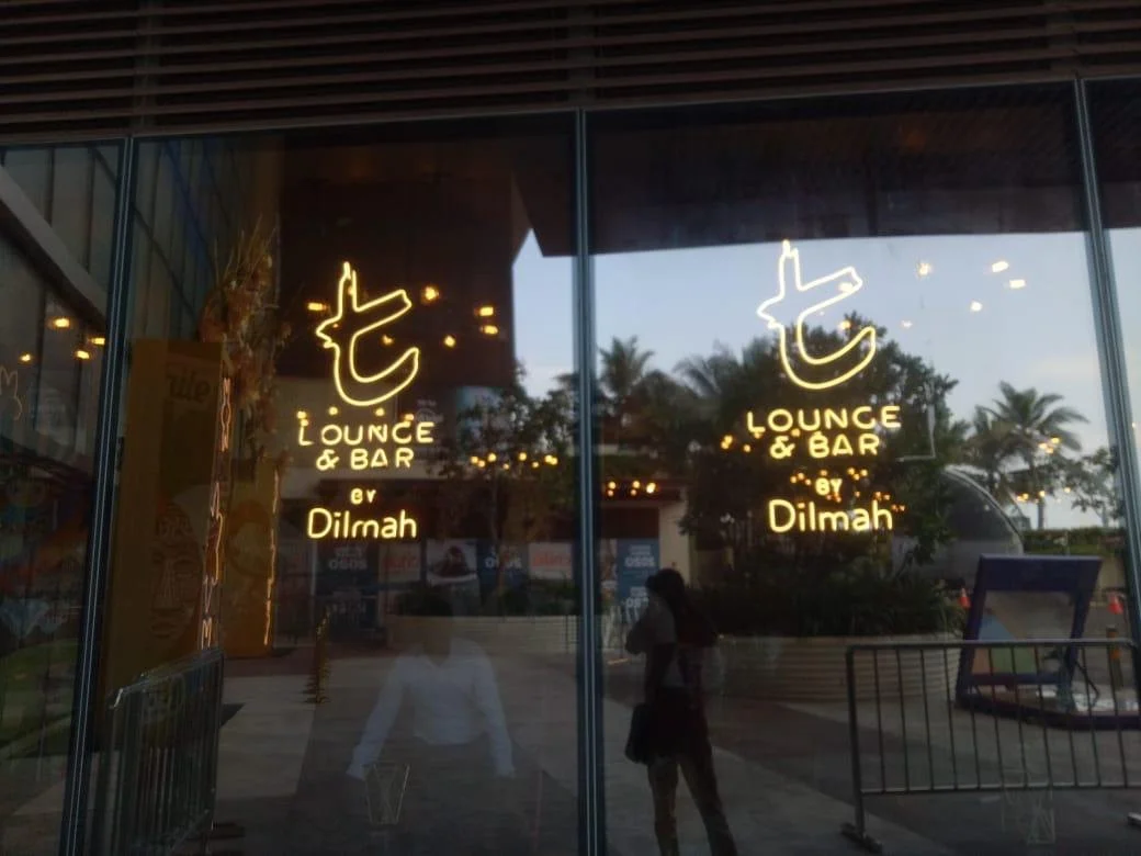 Neon signs on a glass window display the name 'Lounge & Bar by Dilmah' with tropical trees and palm trees outside reflecting on the glass.