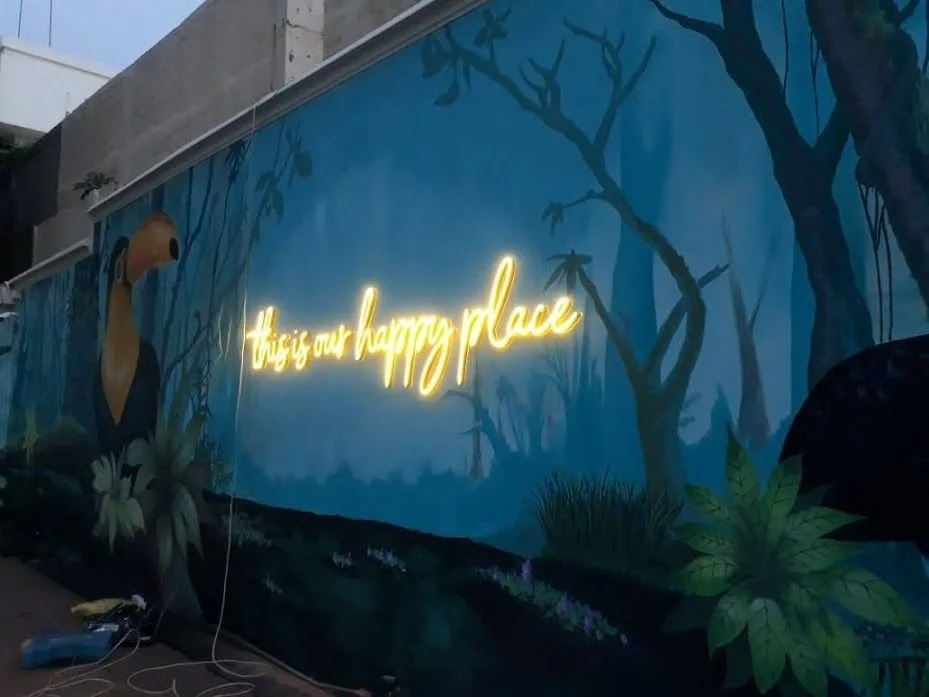 A mural of a jungle scene with trees, plants, and a toucan, with a neon sign that says 'this is our happy place' in yellow cursive lettering.