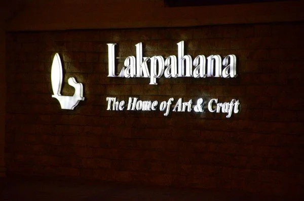Illuminated sign on a wooden wall reads 'Lalkahana The Home of Art & Craft' with a stylized art piece logo to the left.