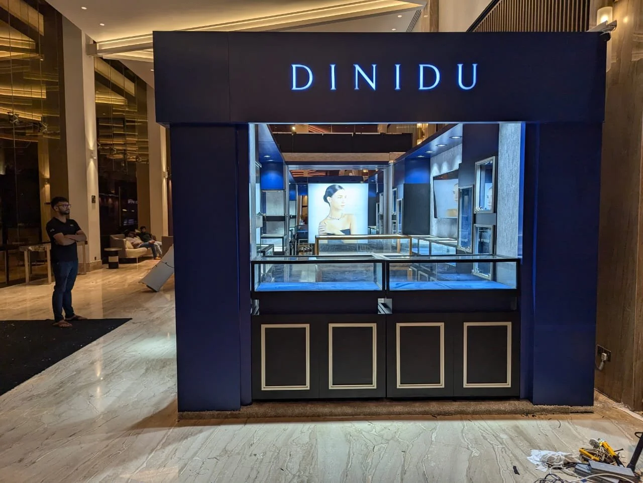 A jewelry kiosk named DINDU located indoors with blue and black decor. Inside the kiosk, glass display cases showcase jewelry, with artwork of a woman on a screen behind.