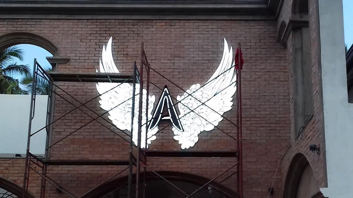 A neon sign with a letter 'A' in the center, flanked by white angel wings, mounted on a brick building with scaffolding in front.