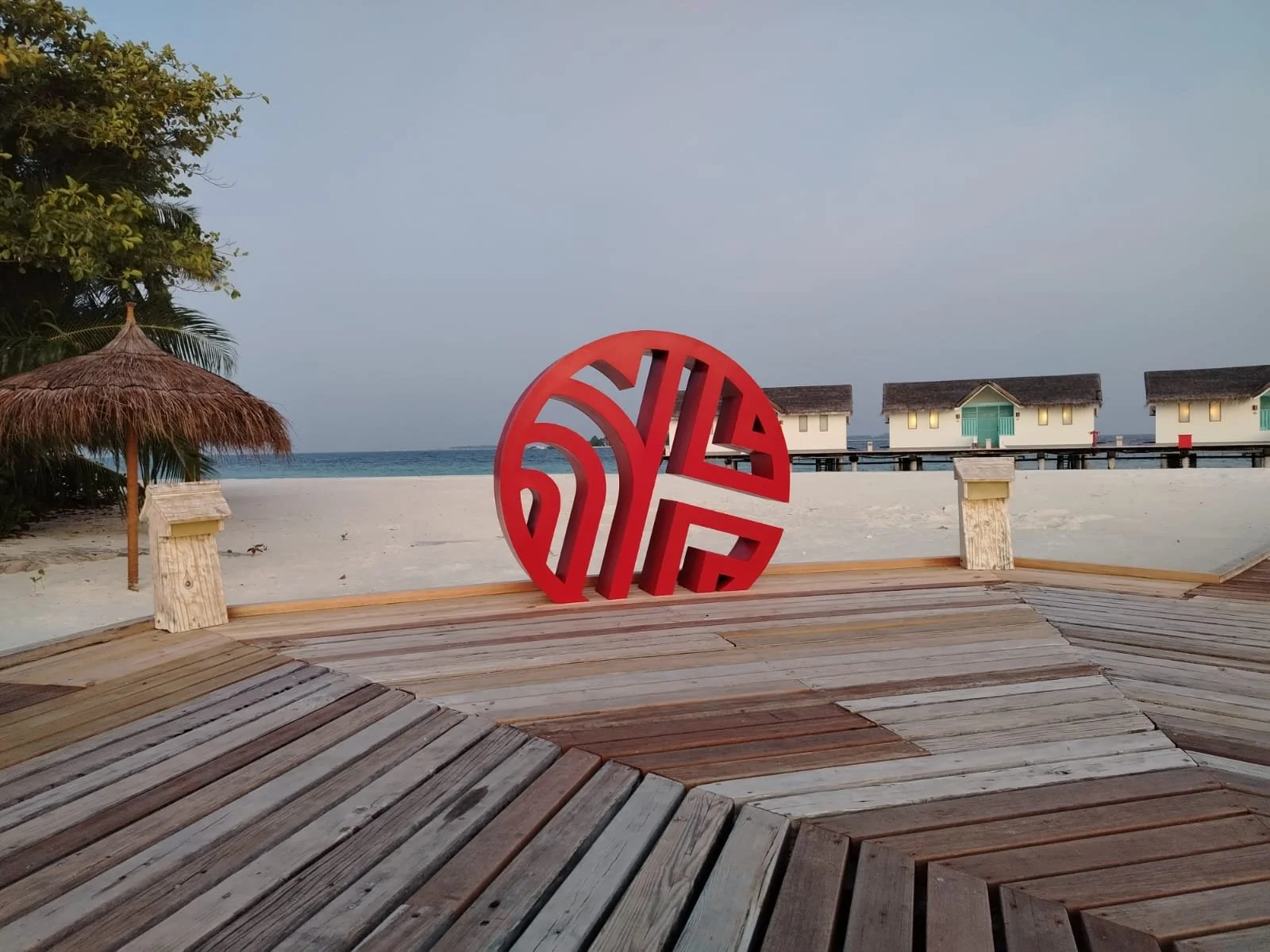 A modern red circular sculpture with intricate cut-out patterns, positioned on a wooden deck at a beachside resort, with overwater bavilions, a thatched umbrella, sand, trees, and the ocean in the background during dusk.