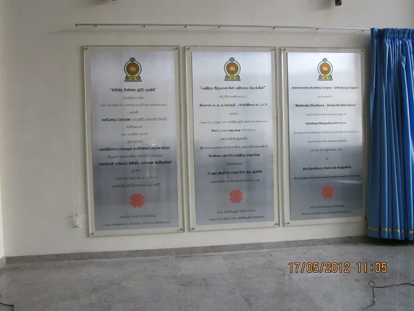 Three official framed documents displayed on a light-colored wall with a shelf above them and blue curtains to the right. The documents have official seals or logos at the top.