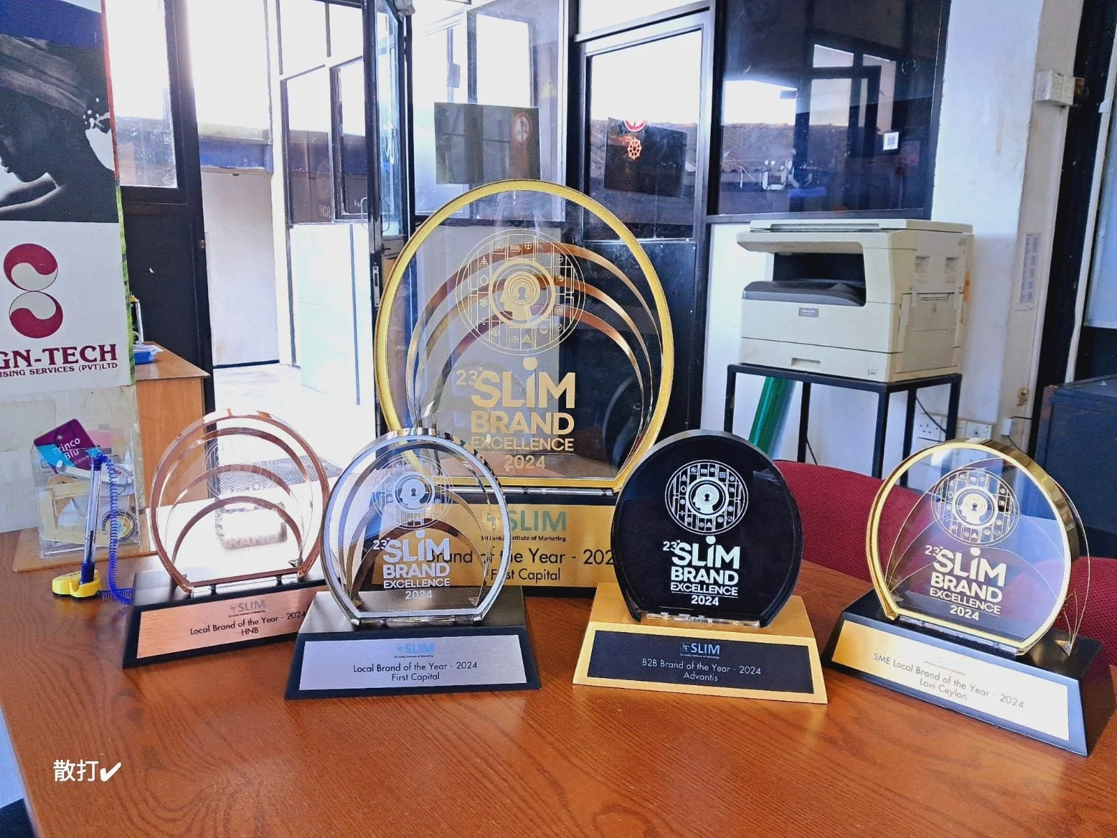 Several awards and plaques on a wooden table, with a large framed award in the background. The awards are for '23'Slim Brand Excellence 2024' in different categories, including Local Brand of the Year, First Capital, B2B Brand of the Year, and SME Lo