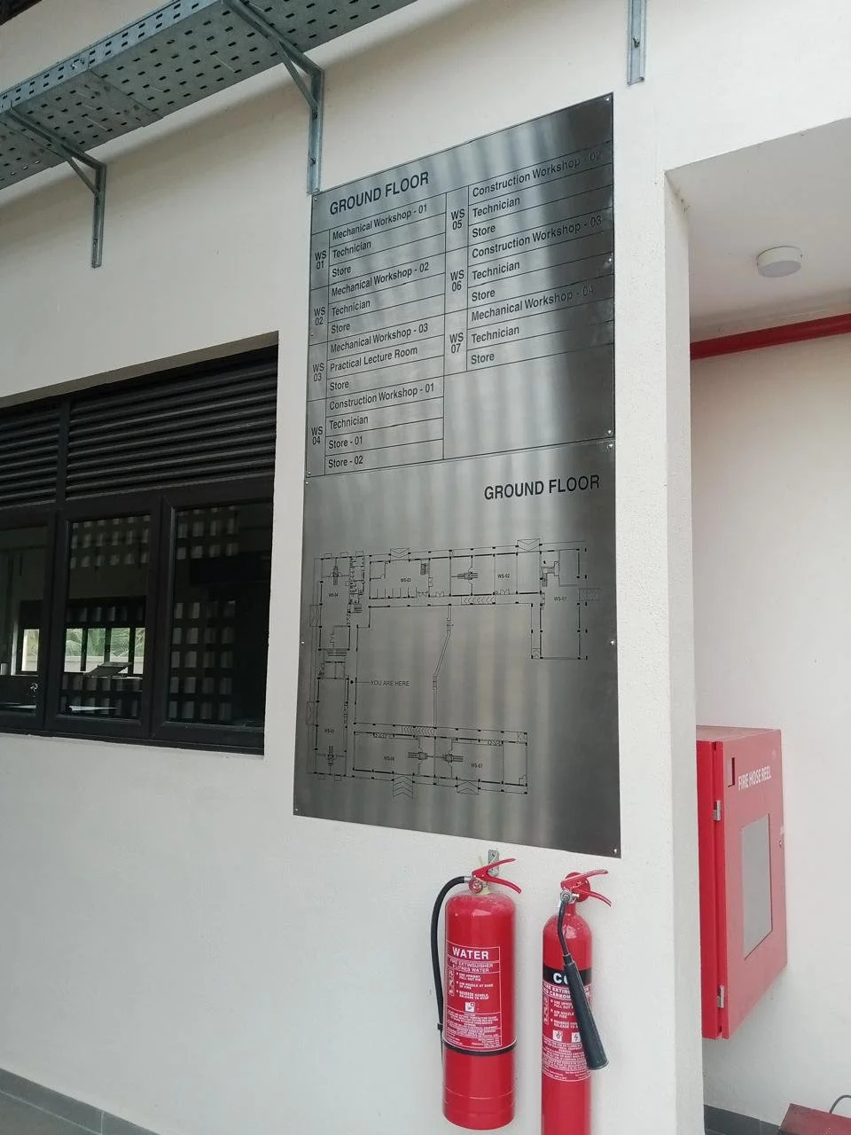 Indoor wall with a black-framed window, a metal directional directory sign, two red fire extinguishers, and a fire hose box.