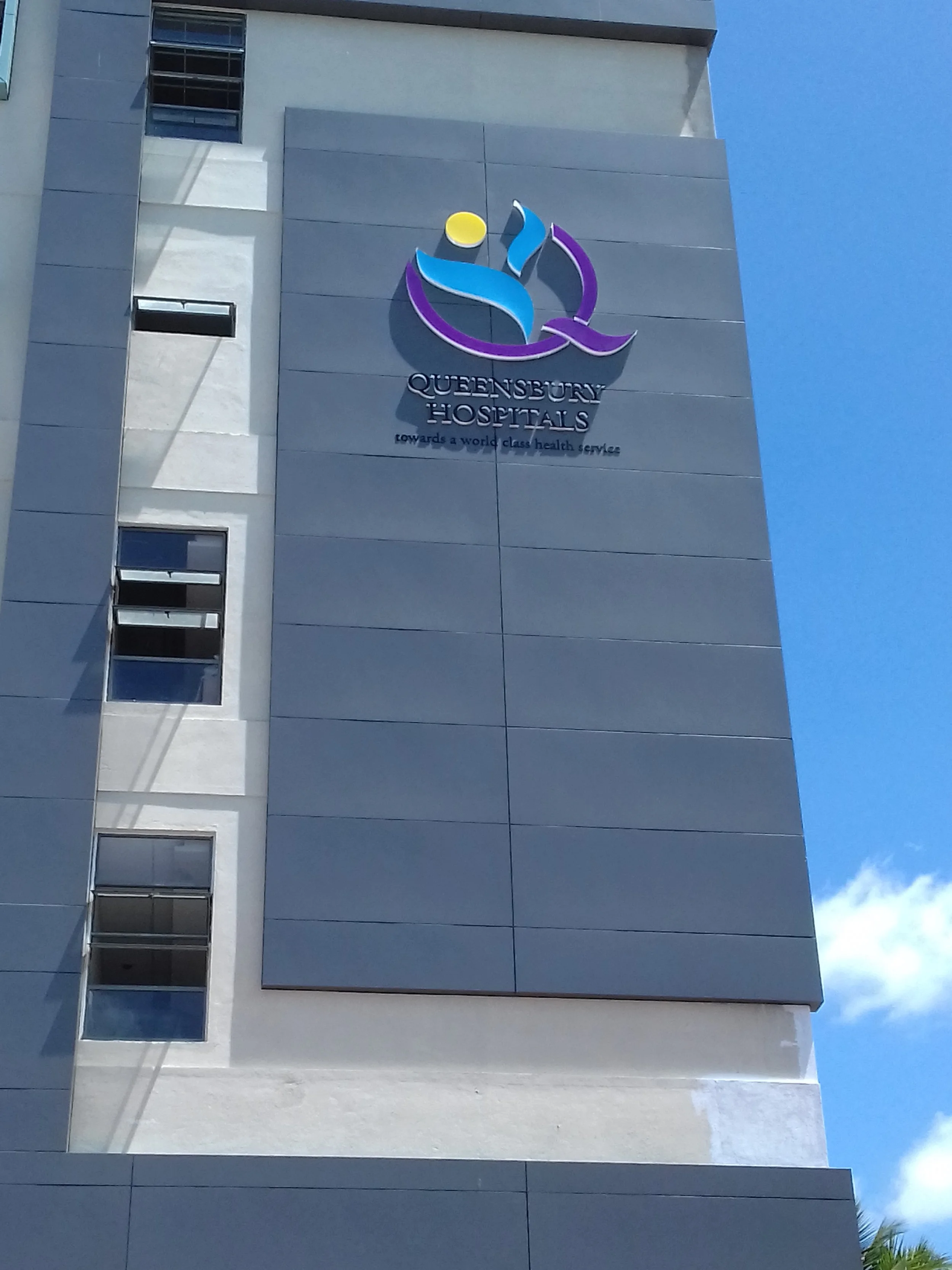 The exterior of Queensbury Hospitals building with a colorful logo featuring a stylized person and a yellow circle, and the text 'Queensbury Hospitals' with the slogan 'towards a world class health service.'
