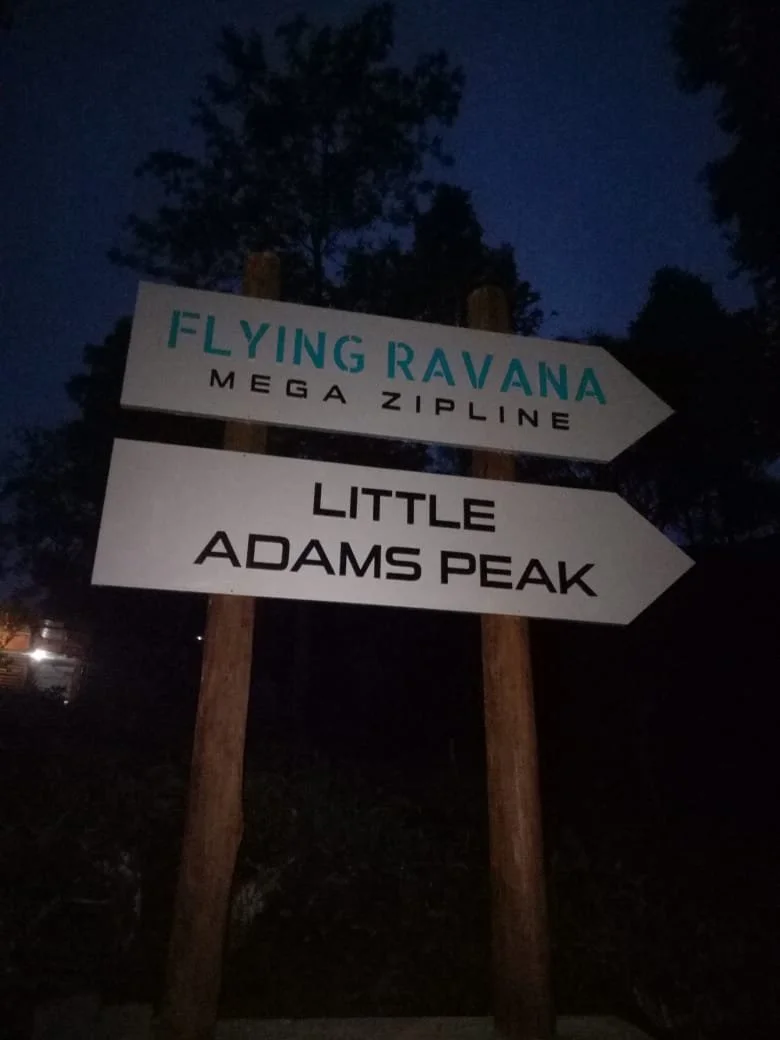 Wooden signpost with four directions near trees at dusk. The top sign reads "Flying Ravana Mega Zipline," the second sign reads "Little Adams Peak," and the third sign reads "Little Adams Peak."