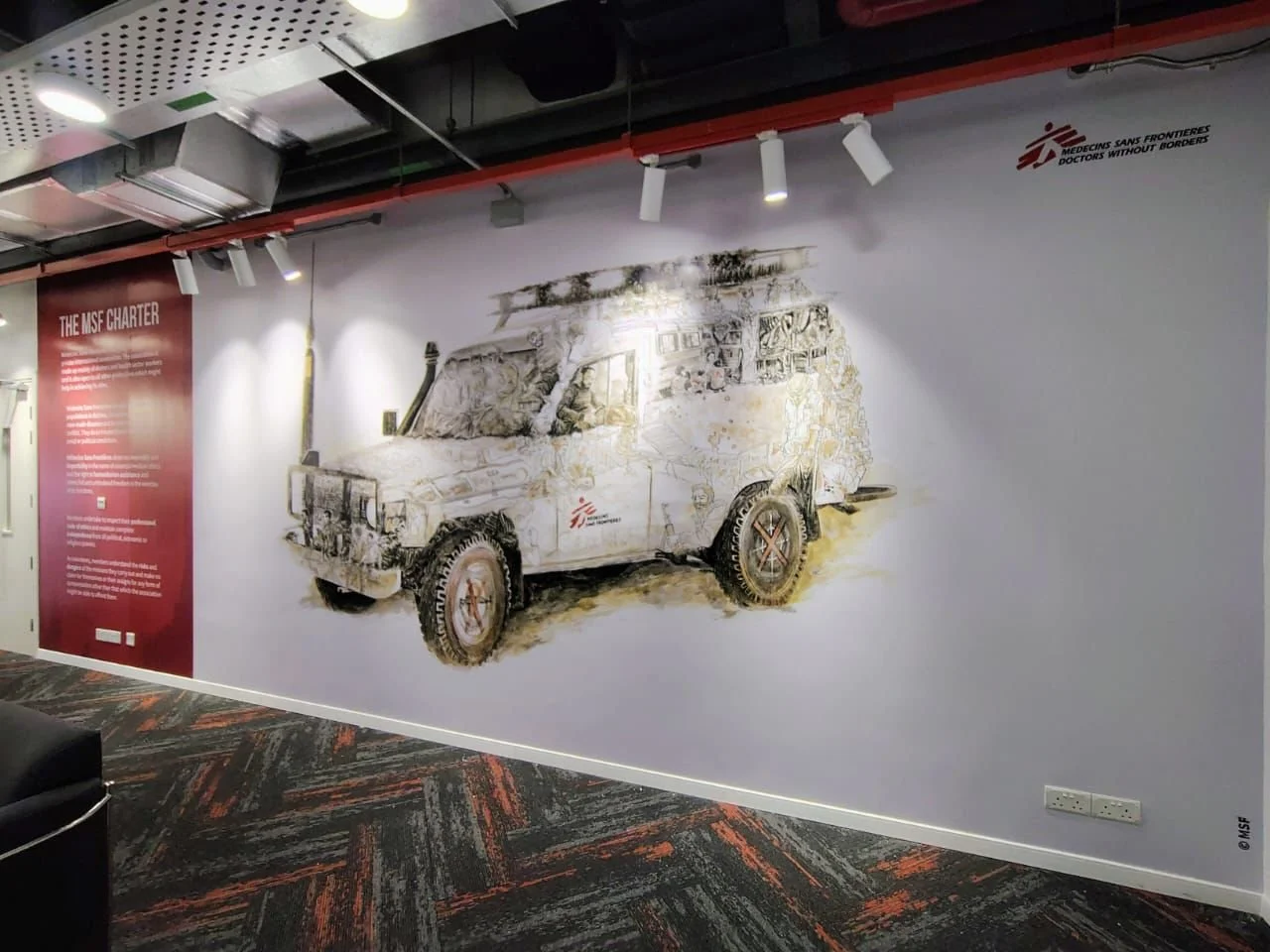 Wall mural of a rugged off-road vehicle with the MSF logo, beneath track lighting, in an indoor space with a patterned carpet and a red information panel.