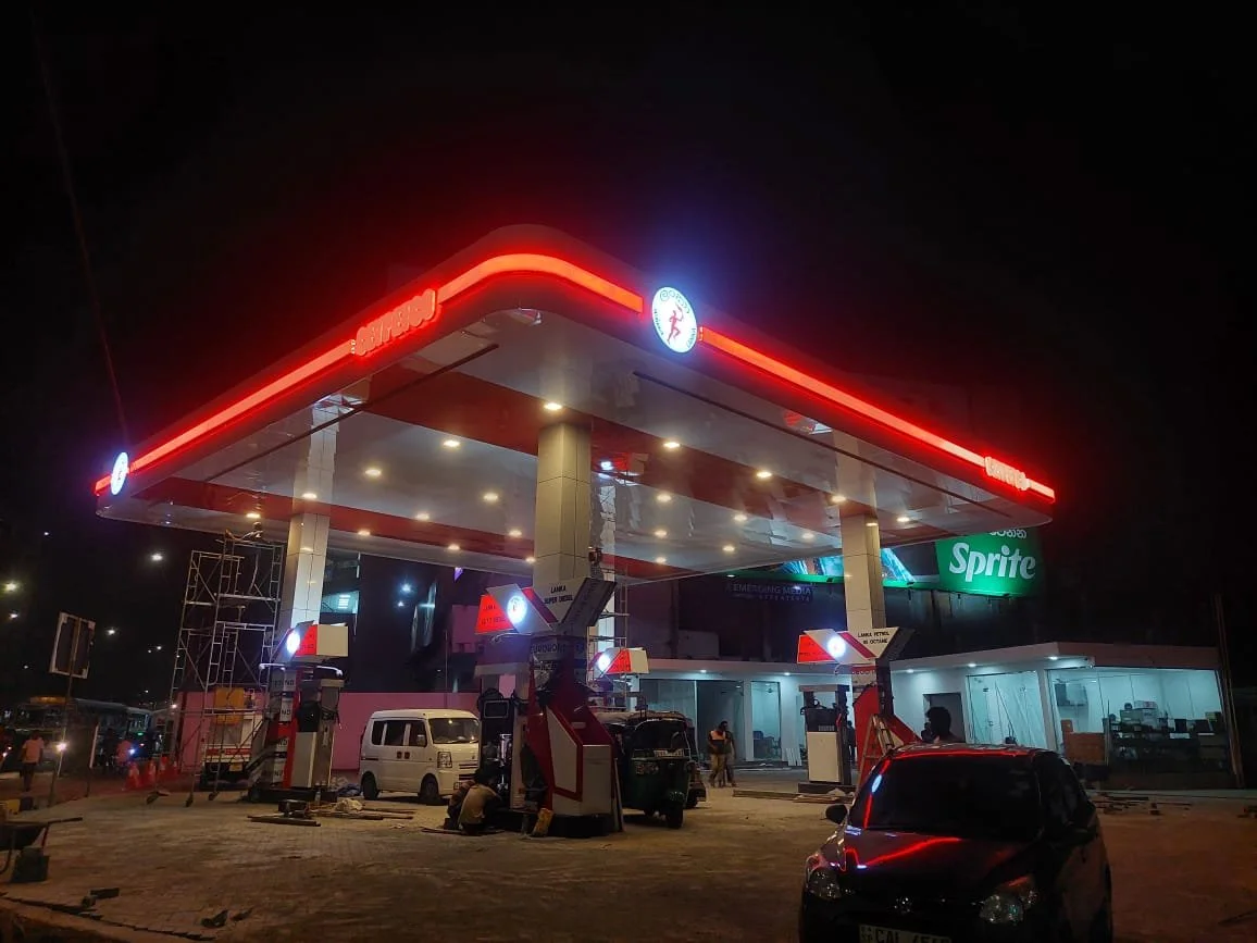 Gas station at night with illuminated signs and fueling pumps, parked vehicles, and a Sprite advertisement.