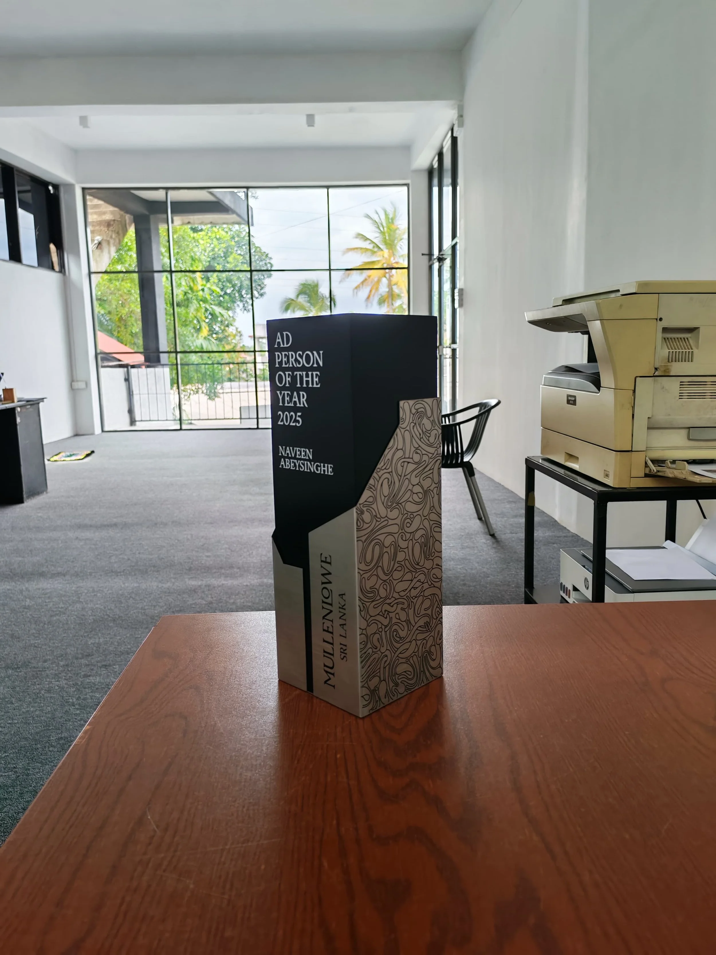 A wooden table with a decorative award or award-like object on it, inscribed with 'Ad Person of the Year 2025' and the name 'Naveen Abeysinghe.' In the background, there is an office environment with a photocopier, a black chair, and large windows sh