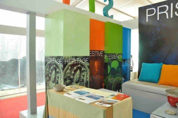 Colorful display with vertical panels labeled HERITAGE, FESTIVE, ESSENCE, and LUSH, with a table in front holding brochures and a bowl, and a sofa with blue and yellow cushions, in a bright indoor space.