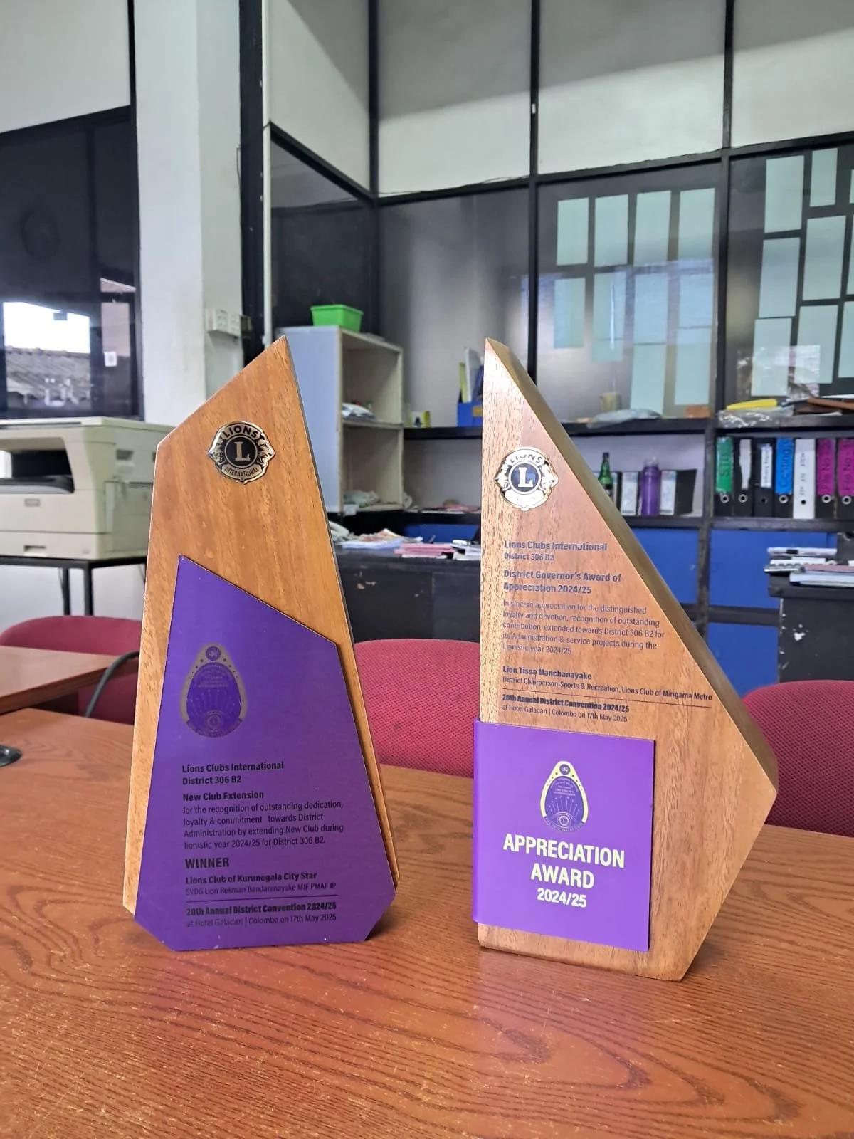 Two wooden trophies on a wooden table, one with purple and one with purple and wooden plaques, in a room with shelves and office equipment in the background.
