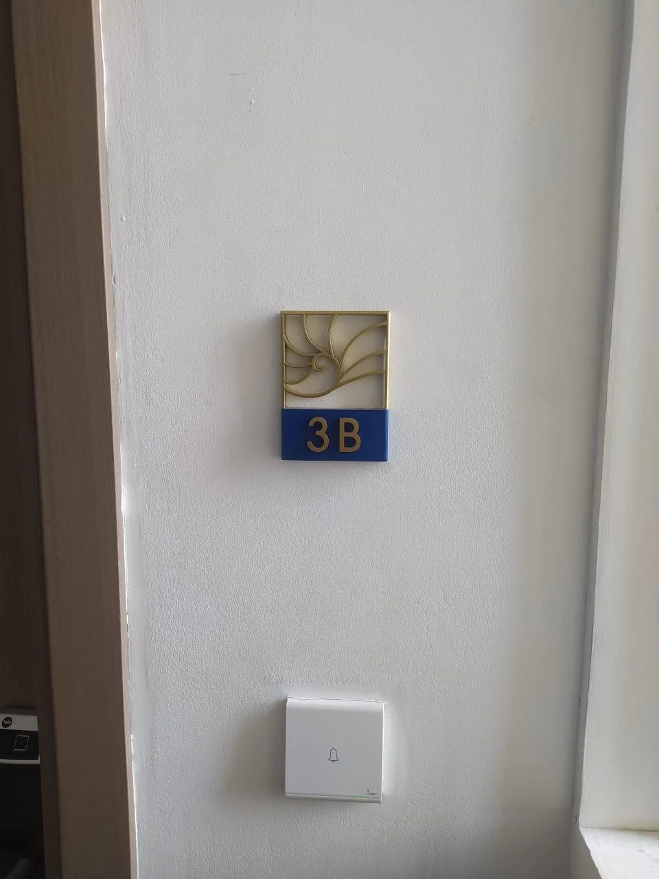 Wall with a hotel room sign labeled 3B, decorative gold-colored wall art above, and a white doorbell with a bell icon below.