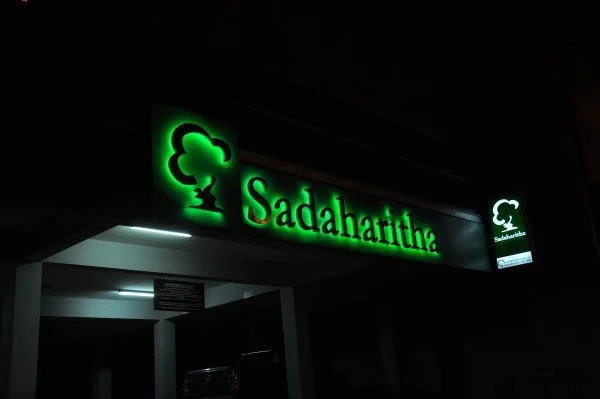 Green illuminated sign for Sadahartha with a logo of a tree on a dark exterior wall.