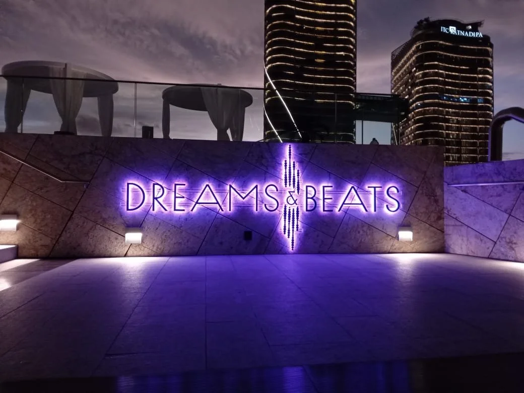 Neon sign reading 'Dreams & Beats' on a tiled wall with city buildings and sky in the background at dusk.