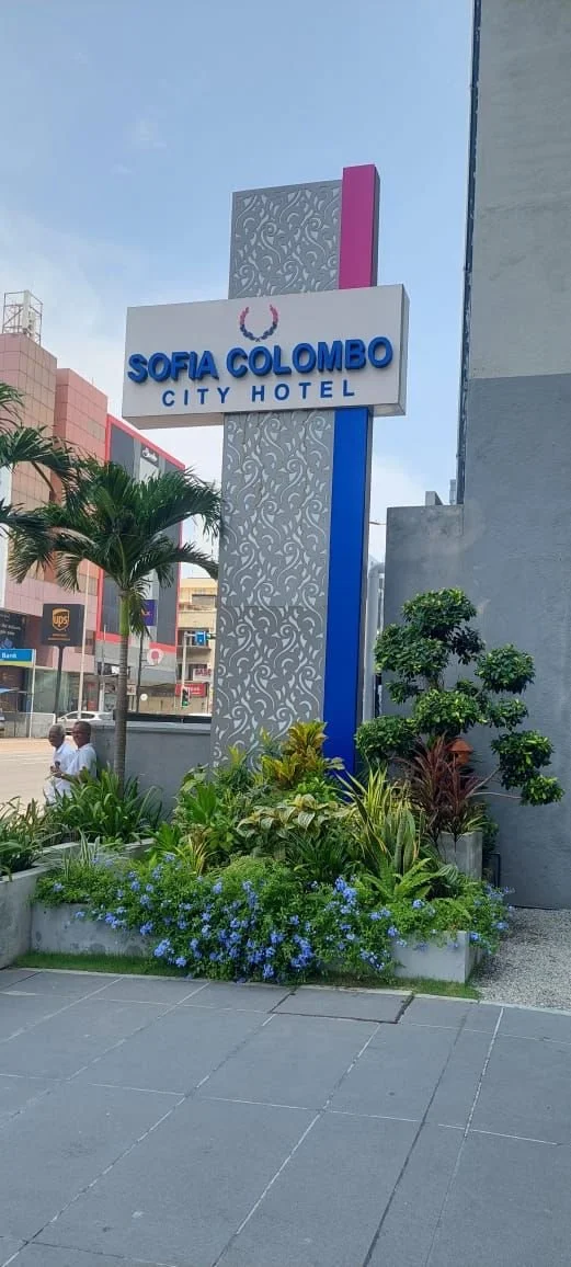 Sign for Sofia Colombo City Hotel on a tall decorative pole, with a palm tree and landscaped plants in front.