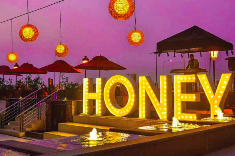 Decorative outdoor setting during dusk with illuminated letters spelling 'HONEY', hanging paper lanterns, umbrellas, and a DJ booth in the background.