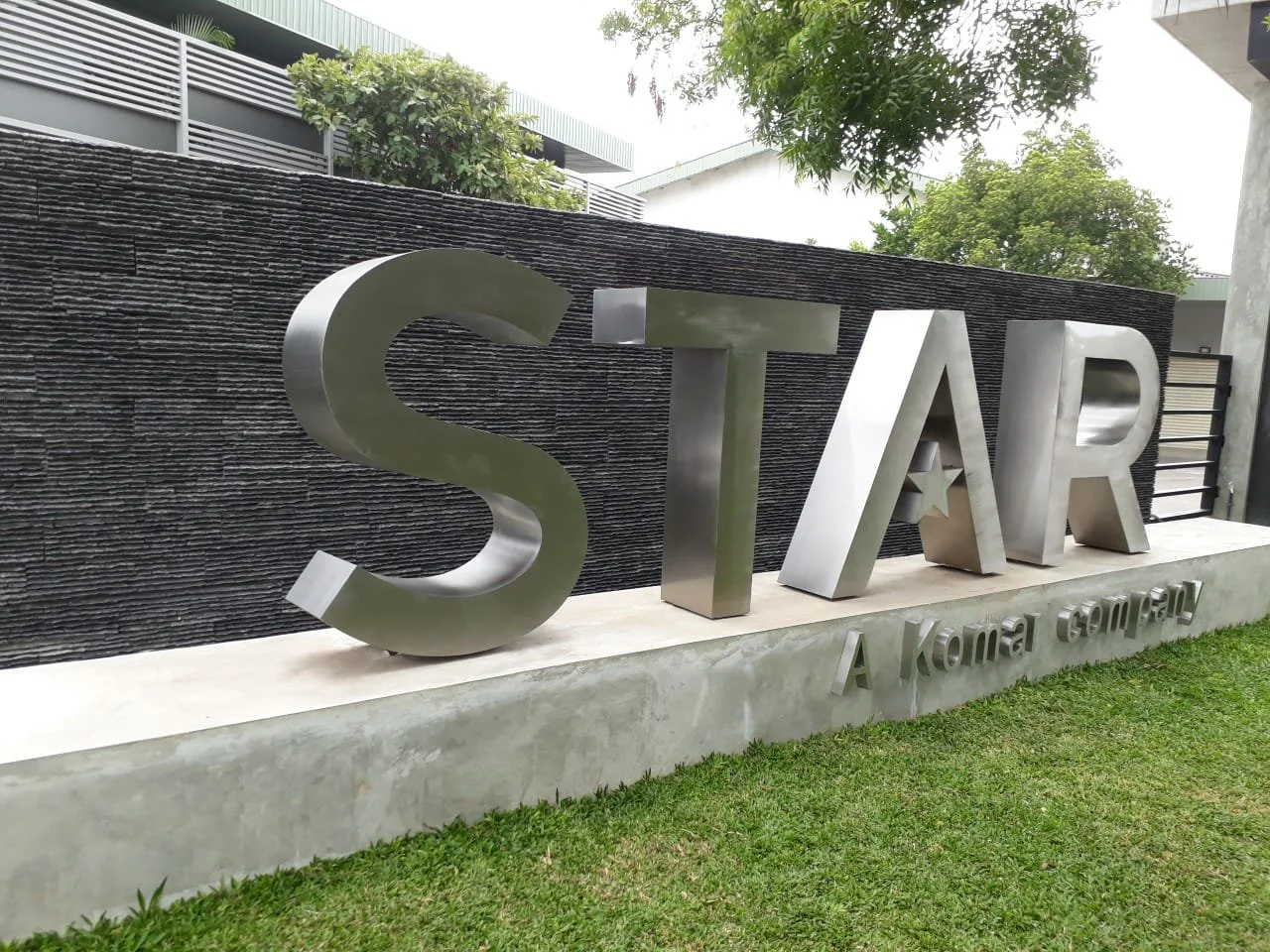 Silver metallic letters spelling STOAR with the phrase "A Kona Company" below on a concrete base in front of a textured dark wall, with trees and buildings in the background.