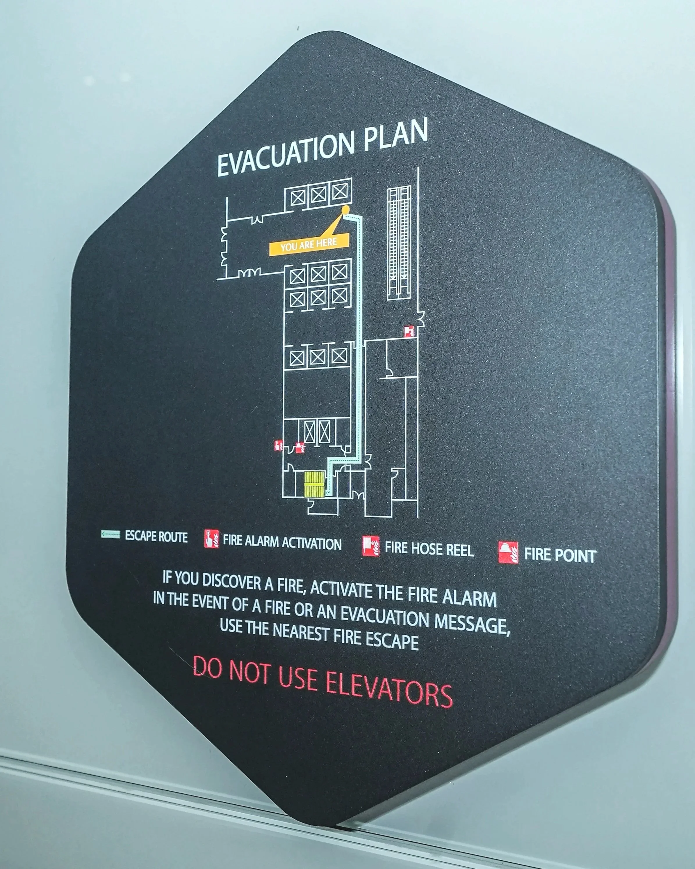 Emergency evacuation plan sign indicating escape route, fire alarm activation, fire hose reel, and fire point with instructions to activate fire alarm and use nearest fire escape in case of fire, and a note not to use elevators.